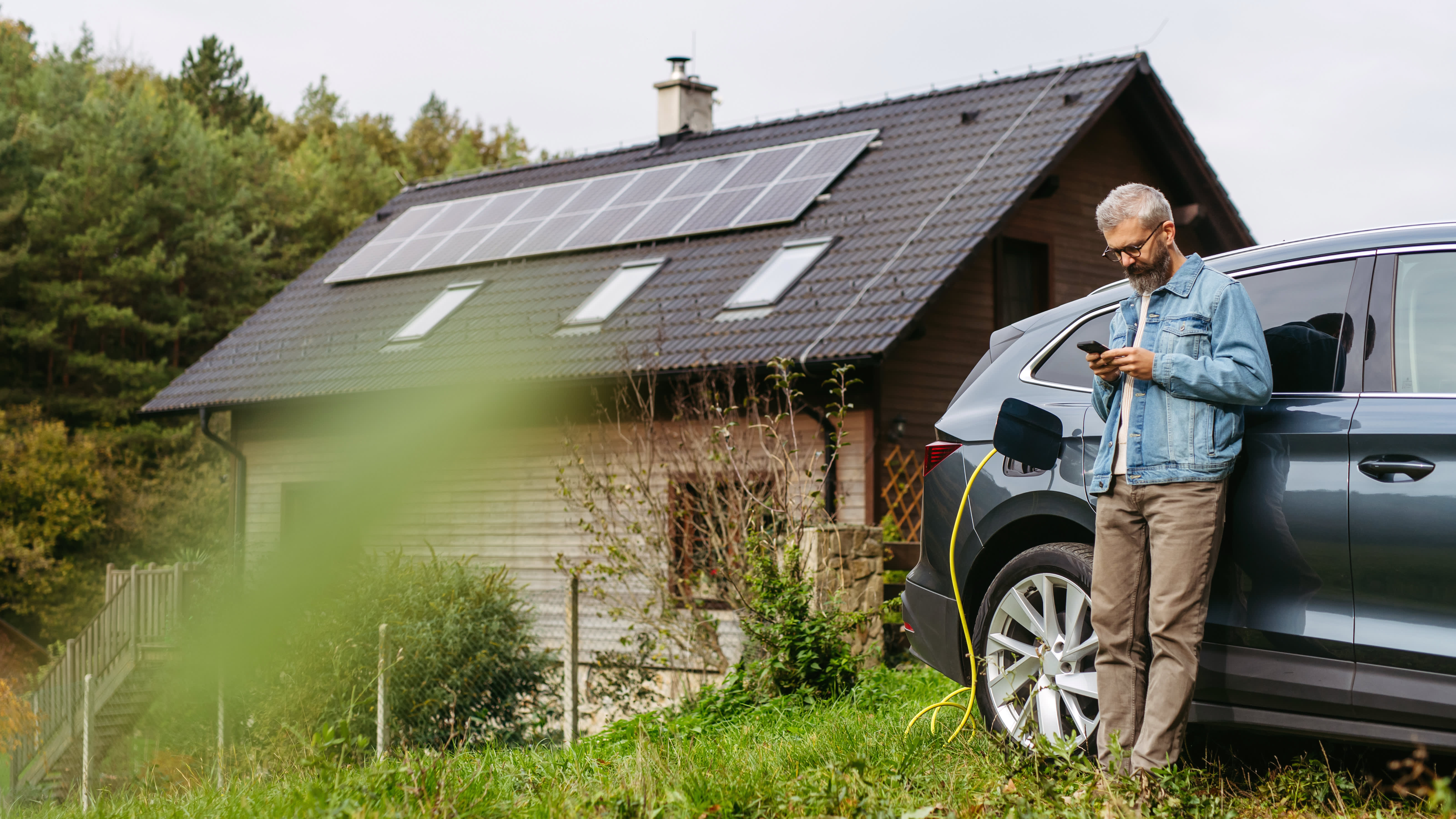 Mann steht mit Smartphone in der Hand an seinem Elektroauto, das gerade lädt, vor einem Haus mit Solarmodulen auf dem Dach
