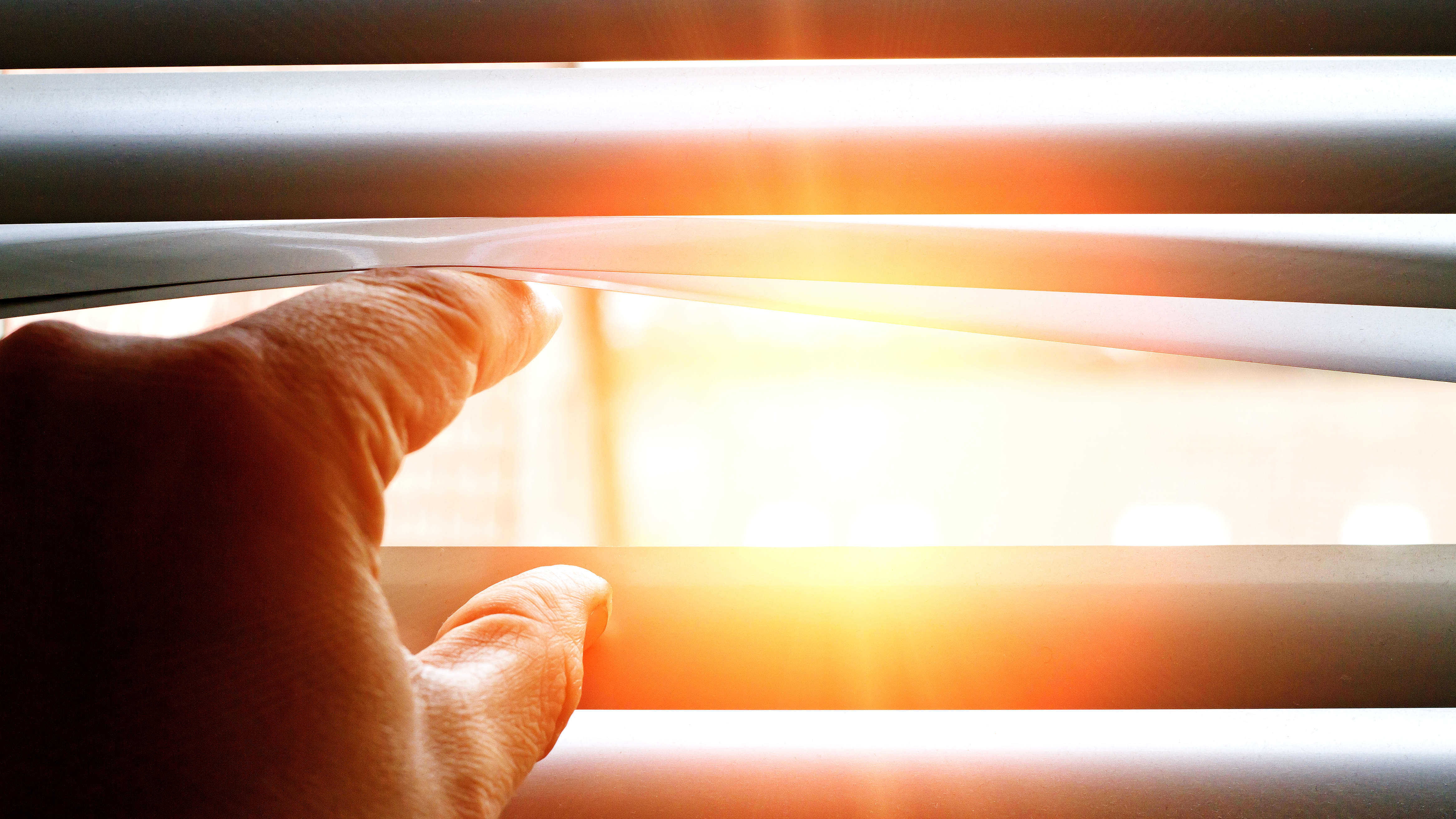 Hand einer Person, die die Lamellen einer Jalousie öffnet, dabei scheint die Sonne scheint durch das Fenster ins Hausinnere.