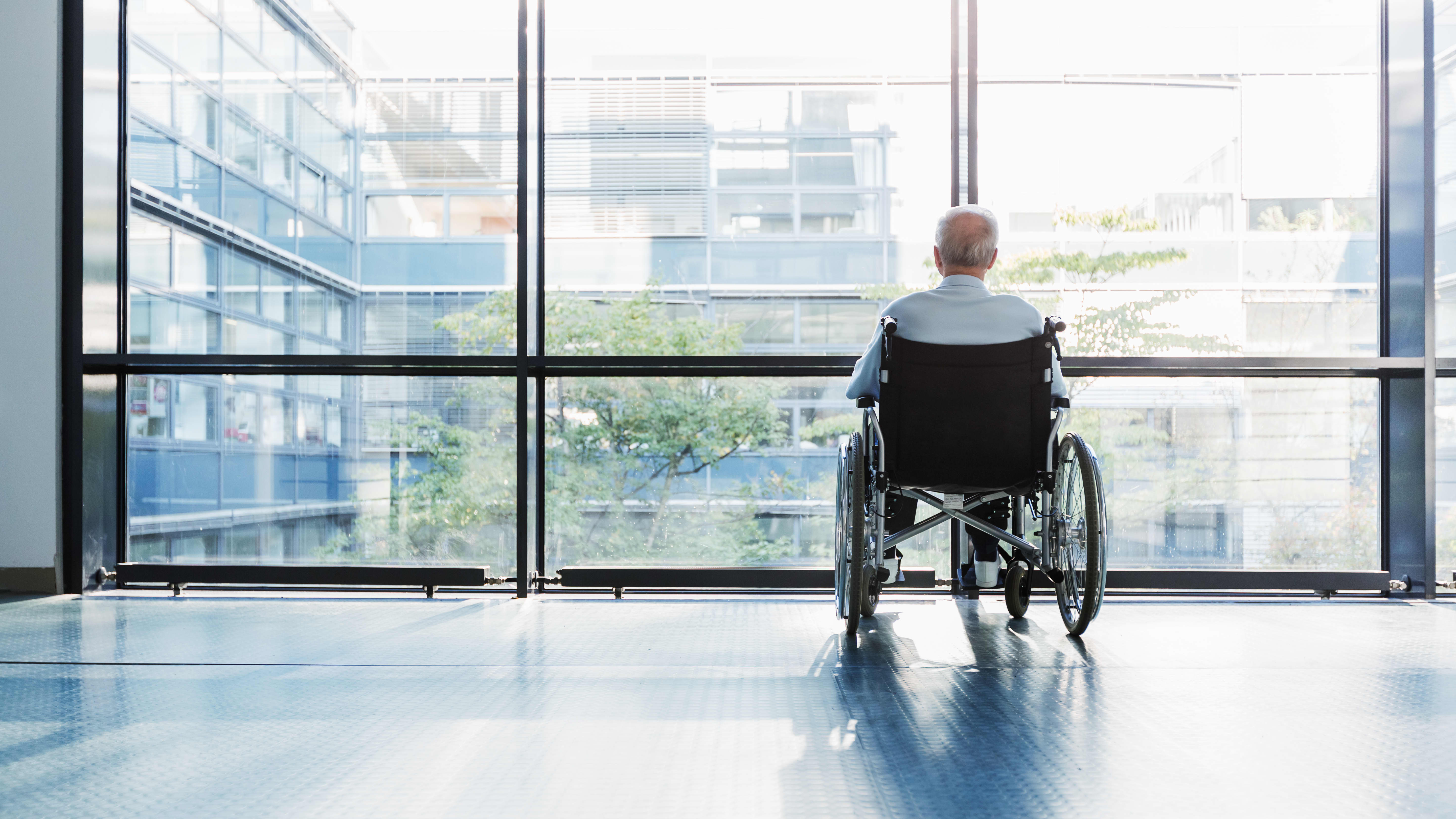 Senior im Rollstuhl blickt aus den bodentiefen Fenstern eines Gebäudes nach draußen