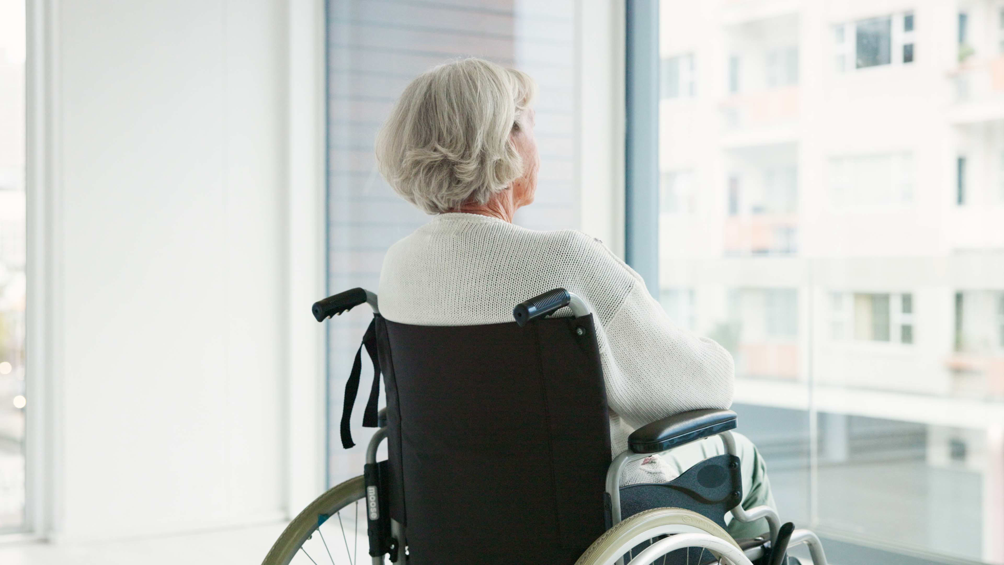 Blick auf eine Seniorin im Rollstuhl, die in einem hellen Raum aus dem Fenster blickt