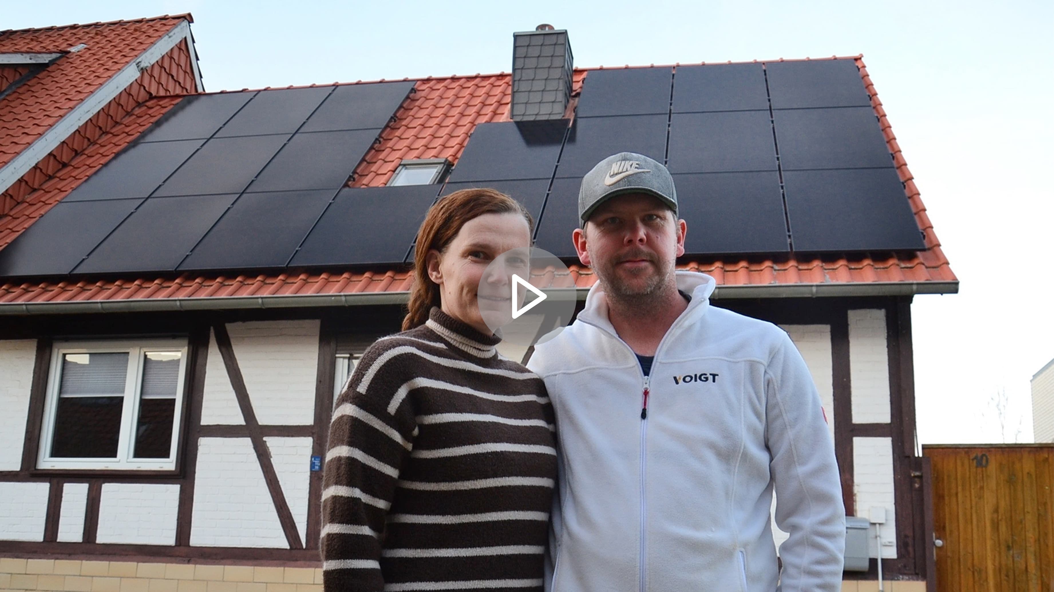 Eine Frau und ein Mann stehen vor einem Haus mit Solarmodulen auf dem Dach. Die Frau lächtelt und trägt einen gestreiften Pullover, der Mann eine weiße Jacke und eine Baseball Kappe. Sie präsentieren ihre neue Solaranlage.