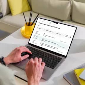 Person arbeitet am Laptop auf einem weißen Tisch mit Smartphone und gelber Vase