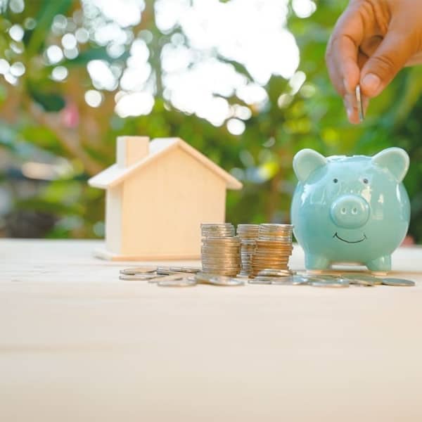 Auf einem Tisch vor grüner Natur befinden sich ein Modellhaus, Münzen und ein grünes Sparschwein.