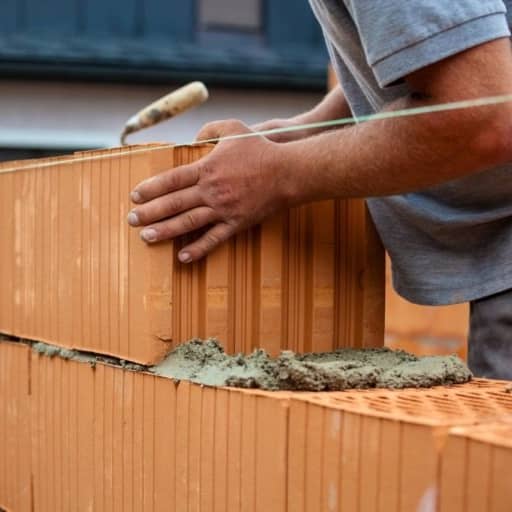 Ein Arbeiter setzt einen weiteren roten Mauerstein auf eine gemauerte Garagenwand.