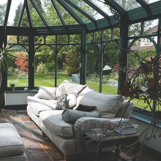 Wintergarten mit Glasdach, schwarzem Stahl-Rahmen, Sitzmöbeln, Steinfliesenboden und Blick in den grünen Garten.