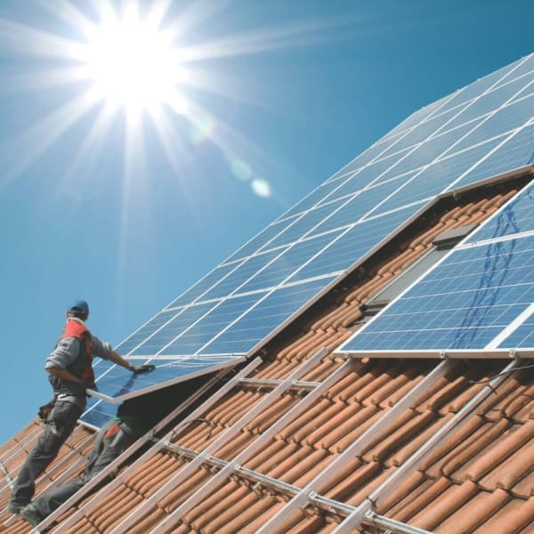 Bauarbeiter stehen vor strahlend blauem Himmel und Sonnenstrahlen auf einem Dach und montieren eine Solaranlage