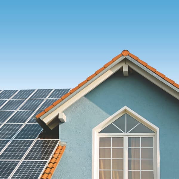 Das Bild zeigt ein Haus mit blauer Fassade und roten Dachziegeln, auf dem mehrere Solarmodule installiert sind. Im Hintergrund ist ein klarer, blauer Himmel zu sehen.