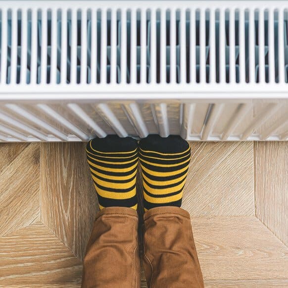 Person mit gelb-schwarz gestreiften Socken wärmt die Füße an einem eingeschalteten Heizkörper, während sie auf einem Holzfußboden sitzt.