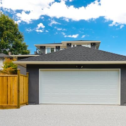 Freistehende moderne Großraumgarage mit einem weißen Doppeltor und dunkler Fassade, umgeben von einem Holzzaun. Im Hintergrund ist ein Wohnhaus und blauer Himmel mit weißen Wolken zu sehen.