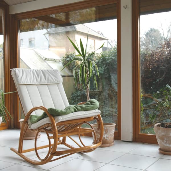 Ein Wintergarten mit großen Holzrahmenfenstern, weiß gefliestem Boden, einem Schaukelstuhl und dekorativen Zimmerpflanzen.