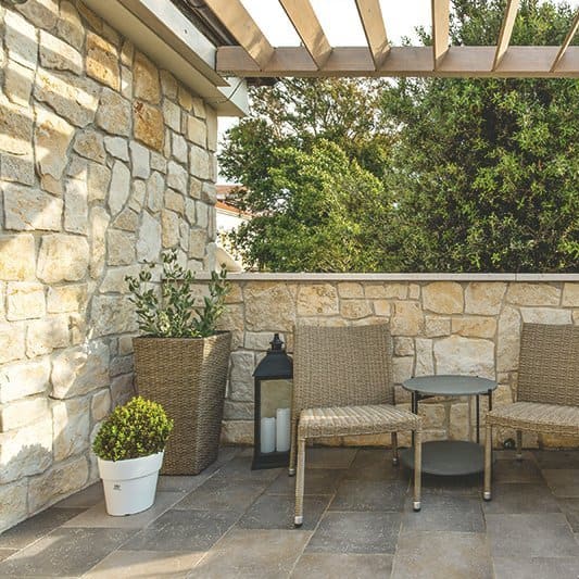 Pergola aus Holz mit festen Lamellen über einer Terrasse mit zwei Stühlen.