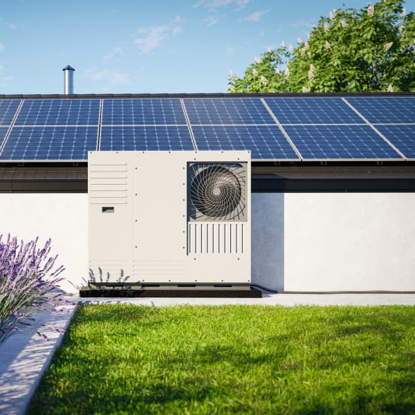 Wärmepumpe vor Haus mit Solaranlage auf dem Dach vor einem Lavendelbeet und grünem Rasen. Im Hintergrund steht ein Windrad