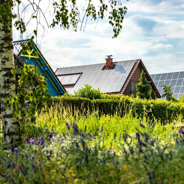 Mehrere Wohnhäuser mit Dächern, auf denen Solaranlagen installiert sind, mit grüner Wiese und Bäumen im Vordergrund unter blauem, leicht bewölktem Himmel
