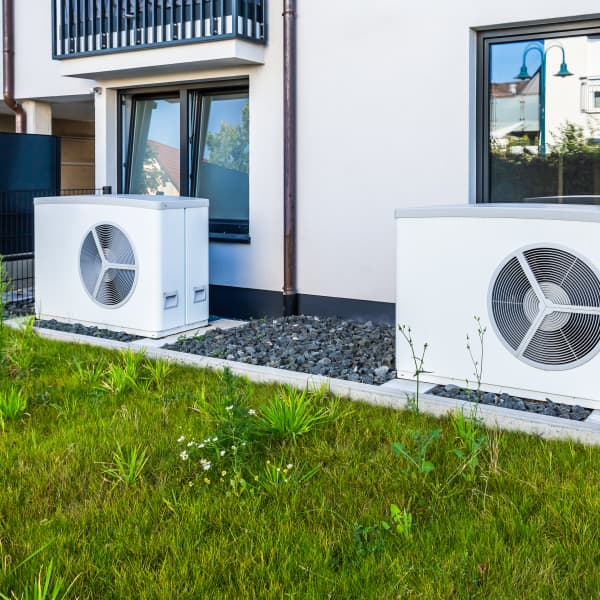 Zwei weiße Wärmepumpen vor weißer Fassade eines Wohngebäudes, davor grünes Gras