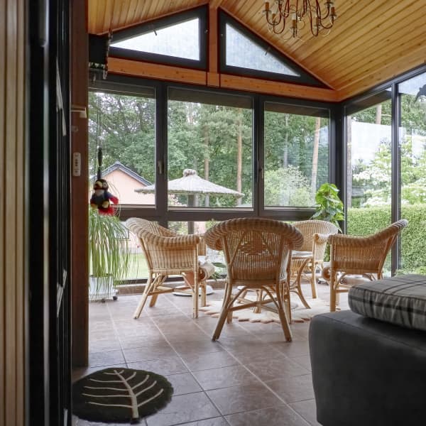 Wintergarten mit Holzdecke, großen Glasfenstern, Rattanmöbeln, Fliesenboden und Blick ins Grüne.