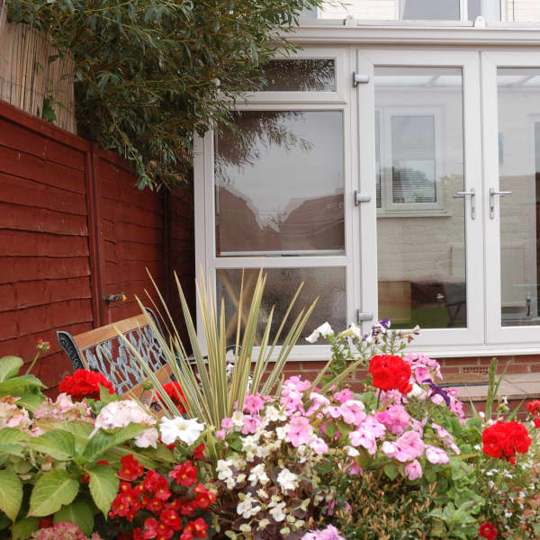 Ein Wintergarten mit großen Glasflächen, umgeben von einem Blumenbeet und Sitzgelegenheiten im Außenbereich.
