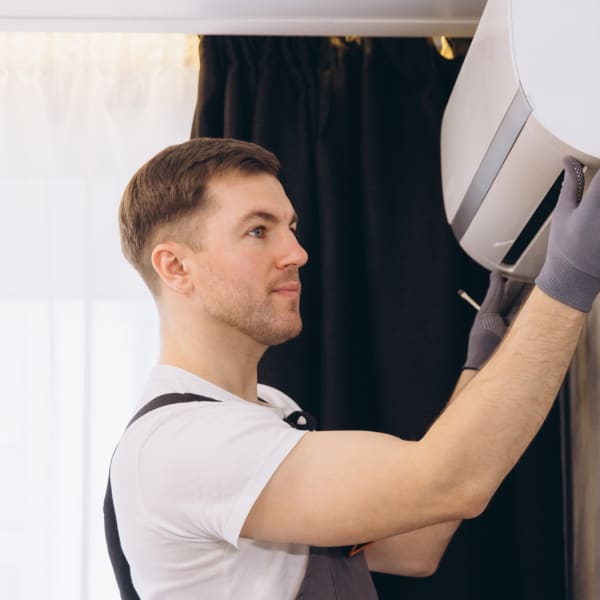 Ein Mann mit Arbeitshandschuhen montiert eine helle, wandhängende Split-Klimaanlage in einem Zimmer; seitliche Nahaufnahme mit Vorhängen im Hintergrund und Teilansicht der Gerätefront.