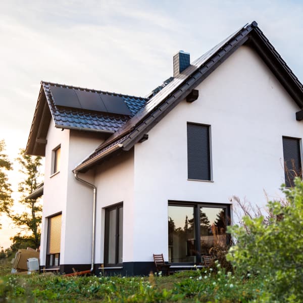 Modernes Einfamilienhaus mit weißer Fassade und bodentiefen Fenstern mit Solarmodulen auf dem Dach steht in einem grünen Garten