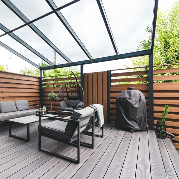 Eine überdachte Terrasse mit  transparentem Glasdach und gemütlicher Lounge-Ecke. Die Terrasse ist mit einem braunen Sichtschutzzaun vom Nachbargrundstück abgetrennt.