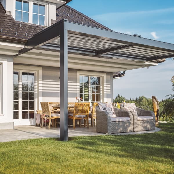 Terrasse eines Einfamilienhauses mit einer Terrassenüberdachung aus Metall. Unter dem Dach stehen gemütliche Rattan-Sessel und ein großer Holztisch mit Stühlen. Der gepflegte Rasen und die umliegende Natur schaffen eine entspannte Atmosphäre.