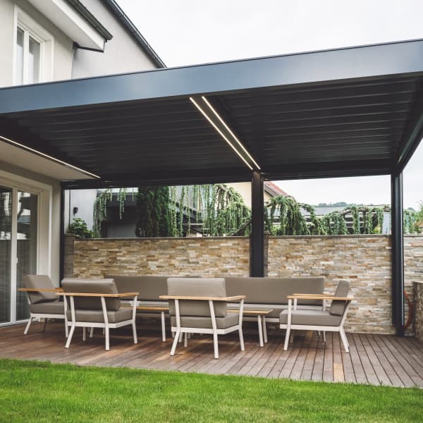Ans Wohnhaus angebaute schwarze Terrassenüberdachung mit Beleuchtung ist über gemütlichen Loungemöbeln platziert.