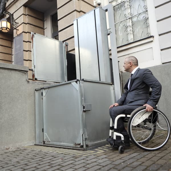 Rollstuhlfahrer vor einem Rollstuhllift vor dem Eingangsbereich eines Gebäudes.