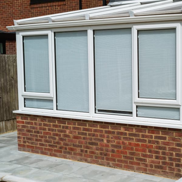Moderner Wintergarten mit weißem Fensterrahmen und integrierten Jalousien, angebaut an ein Ziegelhaus.