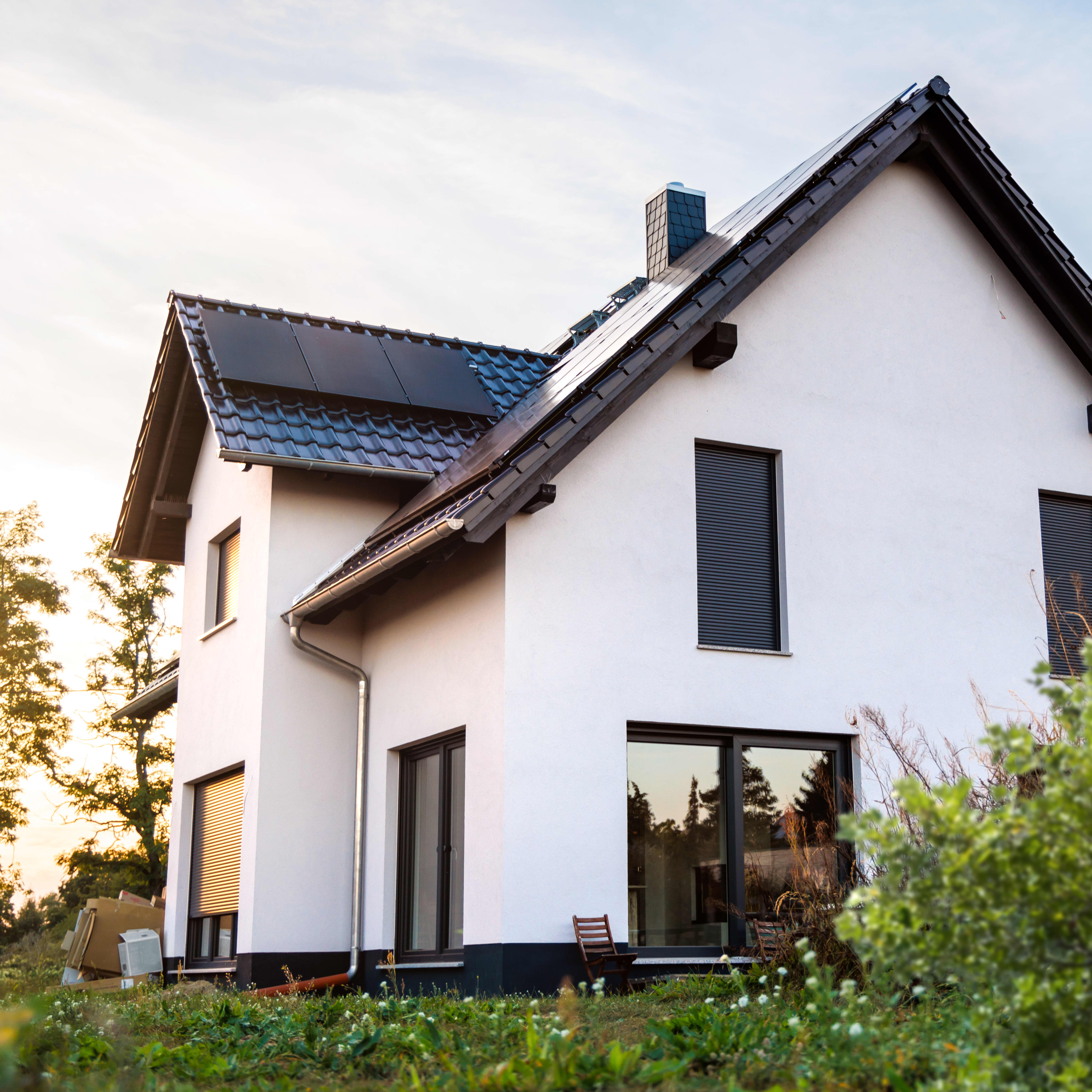 Modernes Einfamilienhaus mit weißer Fassade und bodentiefen Fenstern mit Solarmodulen auf dem Dach steht in einem grünen Garten