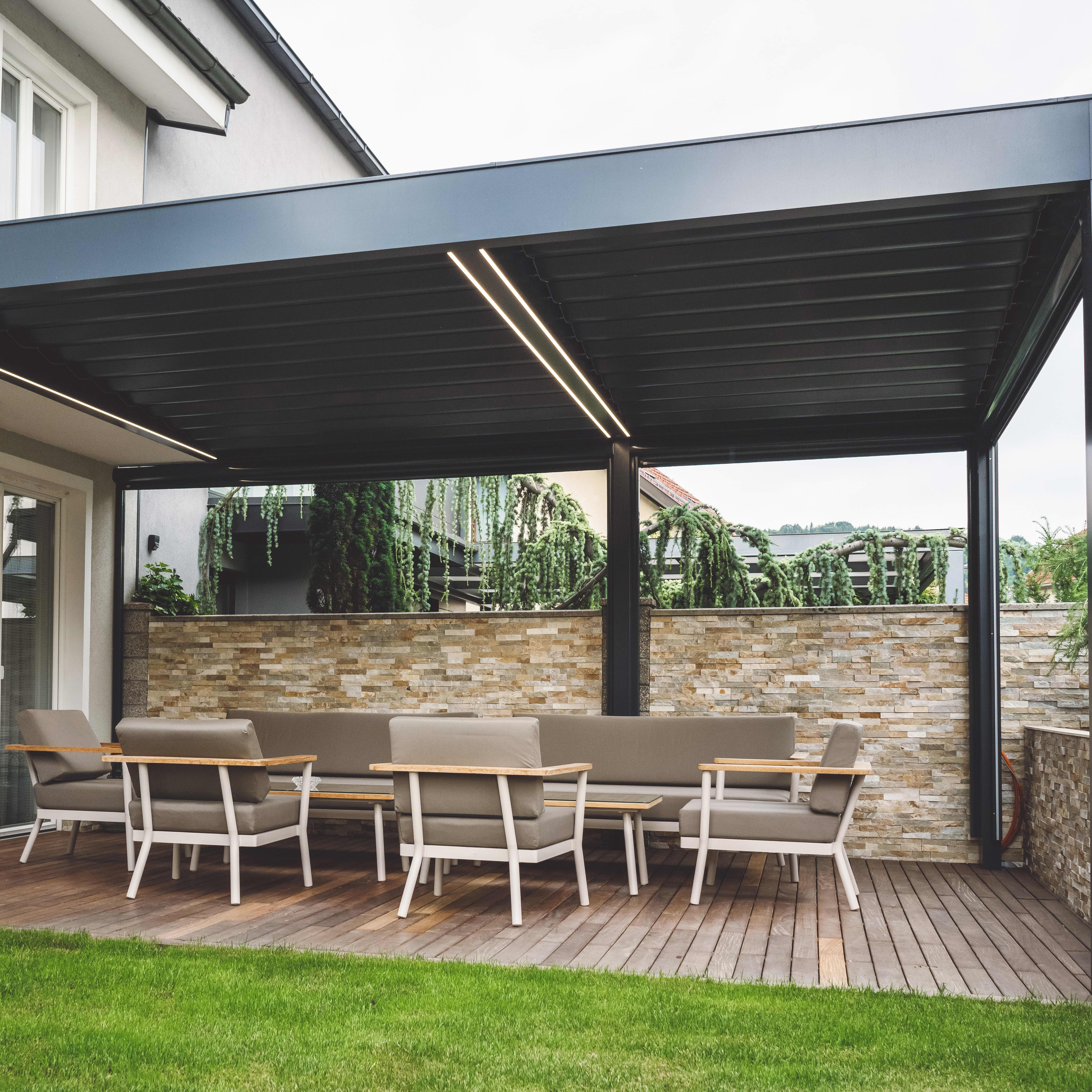 Ans Wohnhaus angebaute schwarze Terrassenüberdachung mit Beleuchtung ist über gemütlichen Loungemöbeln platziert.