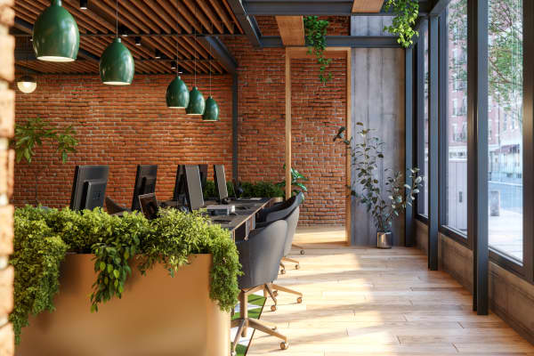 Helles, modernes Büro mit offenen Arbeitsplätzen, Backsteinwand, großen bodentiefen Fenstern und zahlreichen Pflanzen. Stilvolle Pendelleuchten und Holzelemente schaffen eine einladende, natürliche Atmosphäre.