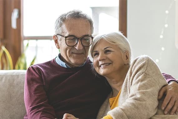 Älteres Paar sitzt Arm in Arm auf dem Sofa in einem hellen Wohnzimmer
