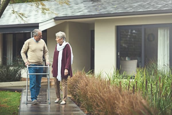 Ein älterer Mann mit Gehhilfe zusammen mit einer Frau vor einem Haus