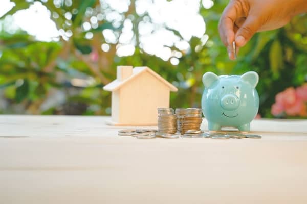 Auf einem Tisch vor grüner Natur befinden sich ein Modellhaus, Münzen und ein grünes Sparschwein.