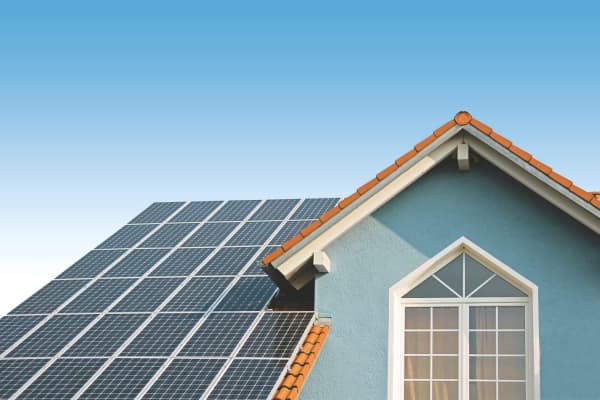 Das Bild zeigt ein Haus mit blauer Fassade und roten Dachziegeln, auf dem mehrere Solarmodule installiert sind. Im Hintergrund ist ein klarer, blauer Himmel zu sehen.