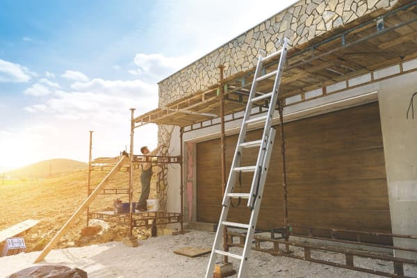Garage mit braunem Tor, an deren Fassade ein Arbeiter Natursteine anbringt. Vor der Garage stehen ein Gerüst und eine Leiter. Die Garagenzufahrt ist noch nicht gepflastert.