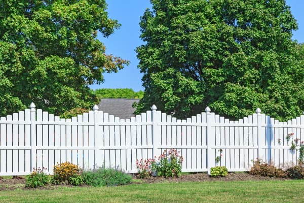 Hoher Zaun aus weißen Latten auf grüner Wiese mit grünen Laubbäumen im Hintergrund