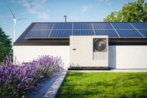 Wärmepumpe vor Haus mit Solaranlage auf dem Dach vor einem Lavendelbeet und grünem Rasen. Im Hintergrund steht ein Windrad