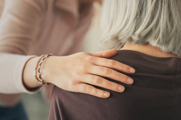 Jüngere Frau legt einer Seniorin mit kurzen grauen Haaren eine Hand auf die Schulter
