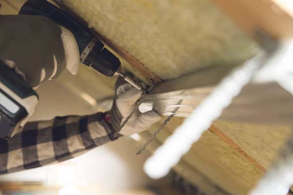 Nahaufnahme der Hände eines Arbeiters, der mit einem Akkuschrauber Bauteile innen an einer Dachschräge befestigt