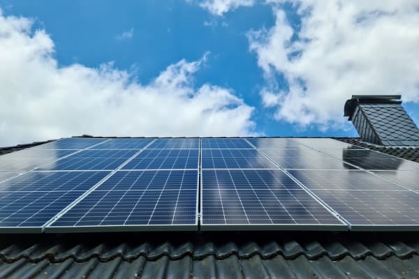 Blick von unten auf ein grau gedecktes Dach mit Schornstein und Solarmodulen bei blauem, leicht bewölktem Himmel