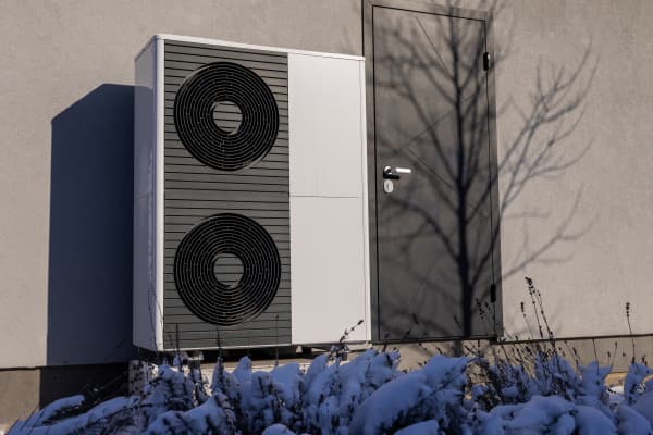 Luftwärmepumpen an der Fassade neben einer Eingangstür mit schneebedeckten Pflanzen im Vordergrund