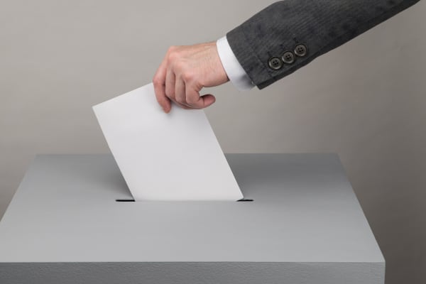 Mann mit Anzug steckt einen Zettel in eine Wahlurne, Aufnahme der Hand und des Arms