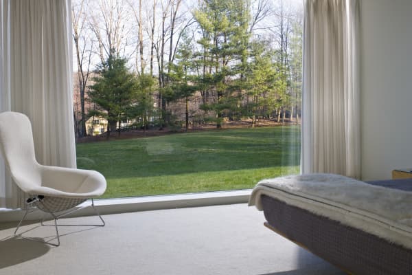 Modernes, minimalistisch eingerichtetes Schlafzimmer mit großem bodentiefem Fenster, hellen Vorhängen, gemütlichem Sessel und Bett mit Felldecke. Draußen: gepflegter Rasen und eine Baumgruppe im Tageslicht.