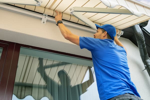 Tageslichtaufnahme an einer Hausfassade: Ein Monteur in blauem Shirt und Kappe arbeitet über einer großen Glas-Schiebetür an einer ausgefahrenen, hellen Gelenkarmmarkise mit Wellenvolant. Sichtbar sind die Metallarme, die Wandhalterungen und die Regenrinne am Dachrand; im Glas spiegelt sich der Arbeitsbereich.