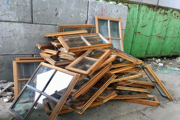 Mehrere braune Fensterrahme aus Holz liegen auf einem Haufen an einer Mauer neben einem Container