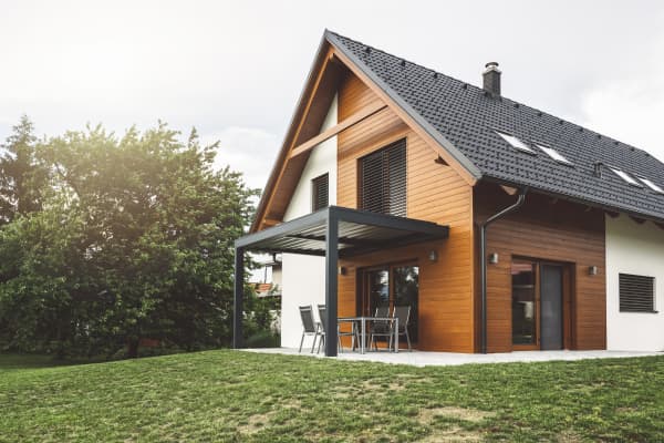 Modernes Einfamilienhaus mit Holzfassade, dunklem Satteldach und einer grauen Terrassenüberdachung im grünen Garten