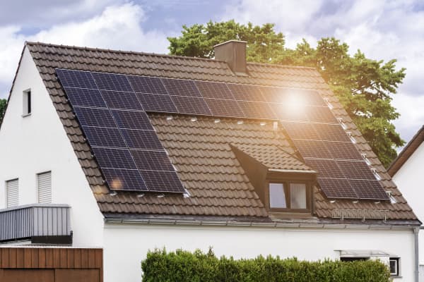 Weißes Einfamilienhaus mit Balkon und Dachgaube, auf dem Dach sind Solarmodule installiert, auf die die Sonne scheint