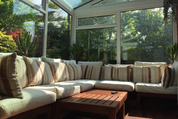 Wintergarten mit großem Ecksofa, Glasdach und Glaswänden, umgeben von Pflanzen und mit Blick auf den grünen Garten.