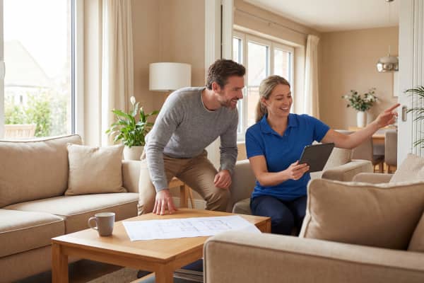 Eine Fachberaterin sitzt mit einem Hausbesitzer im hellen Wohnzimmer und zeigt auf einem Tablet, wo Bewegungsmelder oder Sensoren für die neue Alarmanlage installiert werden können.