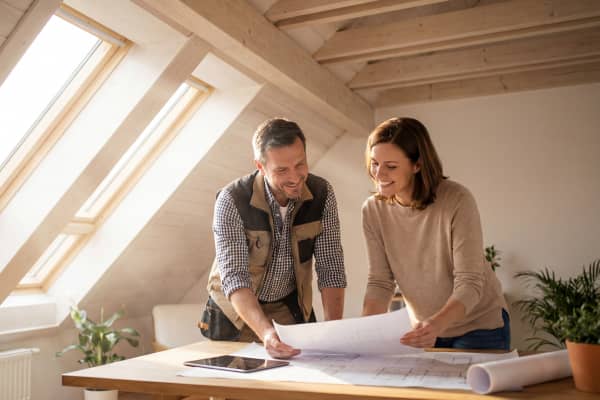 Fachmann und Hausbesitzerin stehen in einem hellen Dachzimmer mit Holzverkleidung und großen Dachfenstern. Beide besprechen gemeinsam Baupläne für den geplanten Fenstereinbau.
